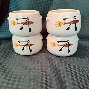 Peut inclure: Quatre bols à soupe en céramique blanc cassé empilés par paires. Chaque bol présente un motif peint à la main d'une fourchette jaune et d'un ustensile marron et noir. Les bols ont de petites poignées de chaque côté.