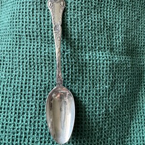 Puede incluir: Una cuchara plateada con un diseño floral ornamentado en el mango. La cuchara tiene un mango largo y delgado y un cuenco redondeado. El mango presenta una parte superior decorativa con un patrón floral y una pequeña inscripción. La cuchara está sobre un fondo verde.