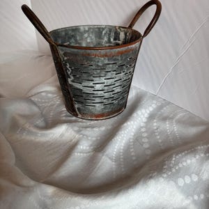 May include: A small, galvanized metal bucket with two arched handles. The bucket has a distressed, rustic appearance with a series of horizontal slots around the sides. The metal has a mottled gray and brown color, suggesting age and use. Ideal for farmhouse decor.