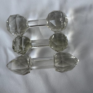 May include: Three clear crystal bottle stoppers with faceted, geometric designs. Each stopper has a round or pointed crystal top connected by a clear stem. The stoppers are displayed on a white surface.