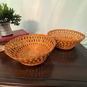 May include: Two woven wicker baskets with a lattice design. The baskets are a warm, light brown color and have a round shape. A small potted plant and books are in the background.