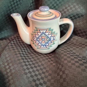 Könnte beinhalten: Eine Keramik-Teekanne mit Deckel, Ausguss und Henkel. Die Teekanne ist cremefarben mit einem geometrischen Design in Blau, Grün und Pfirsich. Der Deckel und der Rand haben blaue und pfirsichfarbene Streifen. Die Teekanne ist zum Aufbrühen von Tee bestimmt.