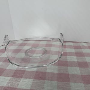 May include: A clear glass serving dish with a shallow oval shape and two decorative handles. The dish has a central raised area and is set on a red and white checkered tablecloth. The background is white.