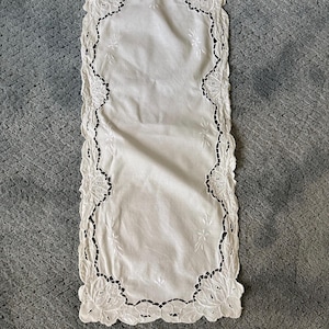 May include: White rectangular table runner with a scalloped edge and floral embroidery. The runner is made of a soft, white fabric and features delicate cutwork around the edges. The design includes embroidered flowers and leaves.