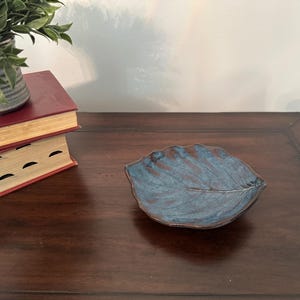 May include: A small, leaf-shaped ceramic dish featuring a glossy glaze with blended shades of deep blue and dark brown. The surface of the dish displays a detailed, embossed vein pattern that mimics the natural structure of a real leaf.