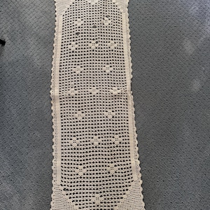 May include: A rectangular off-white crochet table runner featuring an intricate openwork grid pattern with small floral motifs throughout. The runner has a scalloped edge border and a pointed, tapered design at both the top and bottom ends.