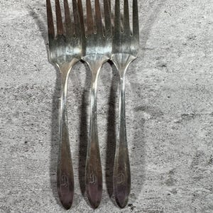 May include: Three silver-toned metal forks arranged side-by-side. Each fork features four long tines and a tapered handle. A decorative letter D is engraved near the base of each handle, showing a consistent design across all three pieces.
