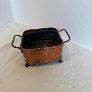 May include: A small, square copper container with two handles and four small feet. The container has a reddish-brown patina with some areas of green oxidation. The interior shows signs of use. A vintage decorative piece.