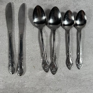 May include: A set of six silver-toned stainless steel utensils consisting of two dinner knives and four spoons. Each piece features a decorative embossed floral pattern on the handle, with a polished finish and a classic, symmetrical design.