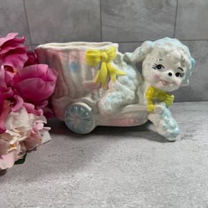 May include: A ceramic planter shaped like a lamb pulling a cart. The lamb is white with blue accents and a yellow bow at its neck. The cart is pink and white with blue floral details and a yellow bow on the side. The piece has a glossy finish.
