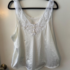 Puede incluir: Una camisola de satén blanco con delicados adornos de encaje a lo largo del escote y los tirantes. La camisola tiene un diseño de cuello en V y está adornada con pequeños botones decorativos. Cuelga de una percha negra contra una puerta blanca.
