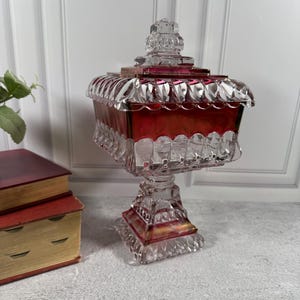 May include: A square glass candy dish with a lid, featuring a deep red center band and clear, faceted edges. The dish stands on a square pedestal base with intricate pressed patterns, showcasing a combination of transparent and ruby-colored glass.