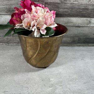 May include: A brass planter with a curved, fluted exterior design and a weathered patina finish. The planter is filled with a bouquet of artificial flowers featuring deep magenta and light pink petals with green leaves.