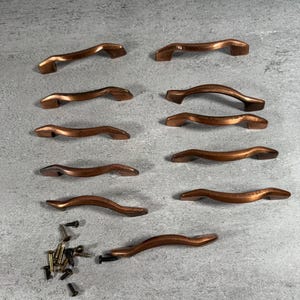 May include: A set of ten curved copper-toned metal cabinet pulls arranged in two columns, featuring a smooth finish and arched design. A small pile of assorted metal screws is included to the left of the bottom handle for installation.