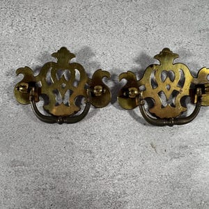 May include: Two matching brass drawer pulls featuring an ornate, openwork backplate with a symmetrical, scroll-like design and a curved, swinging bail handle attached to each side. The metal has a polished, golden-toned finish with visible patina.