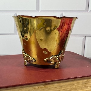 May include: A polished brass planter with a wide, scalloped rim and a tapered body. The base features three ornate, decorative feet with intricate leaf-like detailing. The metallic surface reflects light, showing a warm, golden, and bronze-toned finish.