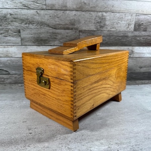 May include: A rectangular wooden shoe shine box with a natural oak finish, featuring finger joint corners, a brass-colored metal latch on the front, and a wooden shoe rest mounted on the top lid. The box is supported by four small wooden feet.