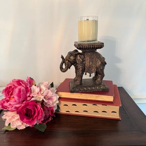 May include: A bronze elephant candle holder with a glass candle sits on two red books. A bouquet of pink and fuchsia flowers rests on a dark wooden surface, creating a decorative display.