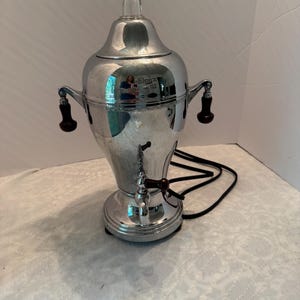 May include: A polished chrome electric percolator with a tall, tapered body, a clear glass finial on the lid, and two dark brown wooden side handles. It features a matching dark brown wooden handle on the front spigot and a black power cord.