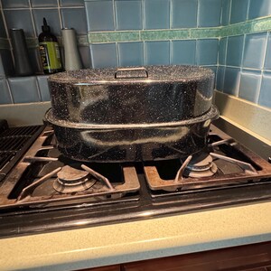 May include: A black speckled enamel roasting pan with a lid, sitting on a gas stove. The pan is rectangular with rounded corners and a handle on the lid. The stove has four burners and is set against a blue tiled backsplash.