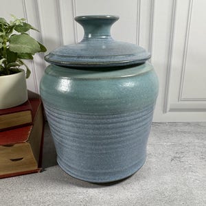 May include: A ceramic canister with a lid, featuring a two-tone glaze with a speckled light blue upper section and a textured, ribbed lower section in a darker blue hue. The lid has a matching blue glaze and a central knob handle for easy removal.