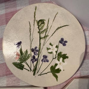 May include: A circular, cream-coloured ceramic coaster with a botanical design. The design features green ferns, purple and white flowers, and blades of grass. The coaster is a decorative item.
