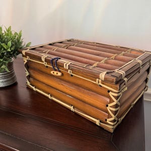 May include: A rectangular storage box constructed from horizontal brown bamboo stalks, featuring light-colored woven reed bindings at the corners and edges. The lid is secured with a small wooden toggle button and a dark blue elastic cord loop.