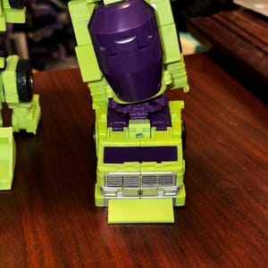 May include: A lime green and purple toy robot with black wheels. The robot has a purple head and a lime green body. The front of the robot has a silver grill and a lime green bumper. The toy is a collectible item.