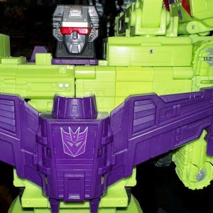 May include: A close-up of a green and purple toy robot. The robot has a silver head with red eyes and a purple chest with a symbol. The robot's body is primarily green with purple accents.