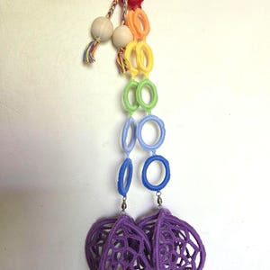 Peut inclure: Un mobile suspendu composé d'une chaîne verticale d'anneaux recouverts de tissu aux couleurs de l'arc-en-ciel, incluant rouge, orange, jaune, vert, bleu clair et bleu foncé. Deux perles en bois naturel avec des pompons multicolores pendent du haut, se terminant par deux sphères en crochet violet.
