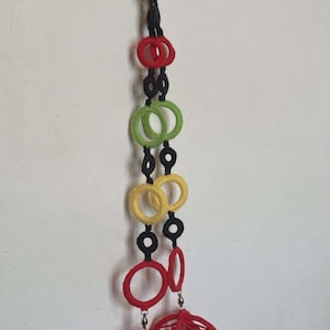 Puede incluir: Una decoración colgante hecha a mano y colorida. Presenta una serie de anillos envueltos en hilo verde, amarillo, rojo y negro, conectados por hilo negro. Dos esferas rojas caladas cuelgan en la parte inferior. La decoración está suspendida contra una pared blanca.
