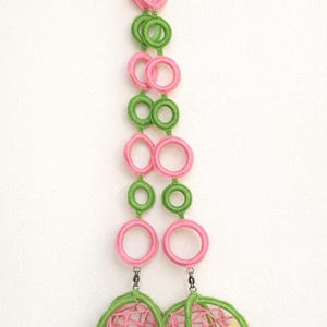 Può includere: Una decorazione da parete verticale con una serie di anelli all'uncinetto alternati rosa e verdi collegati in due colonne. Alla base, due strutture sferiche a gabbia realizzate con filo verde e rosa contengono piccoli accenti floreali gialli.