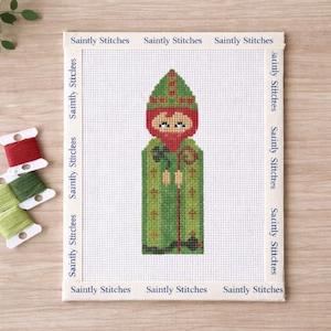 May include: Cross-stitch of a leprechaun wearing a green hat and coat, with a red beard and face. The design is on a white canvas, framed by a beige border with the text "Saintly Stitches". Spools of red and green thread are in the corner.