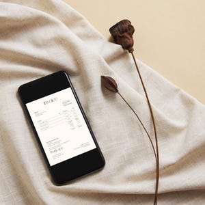 May include: A black smartphone displays an invoice on a cream-colored screen, resting on a beige linen cloth. Dried seed pods and stems add a natural element to the composition. The word "Invoice" is visible.