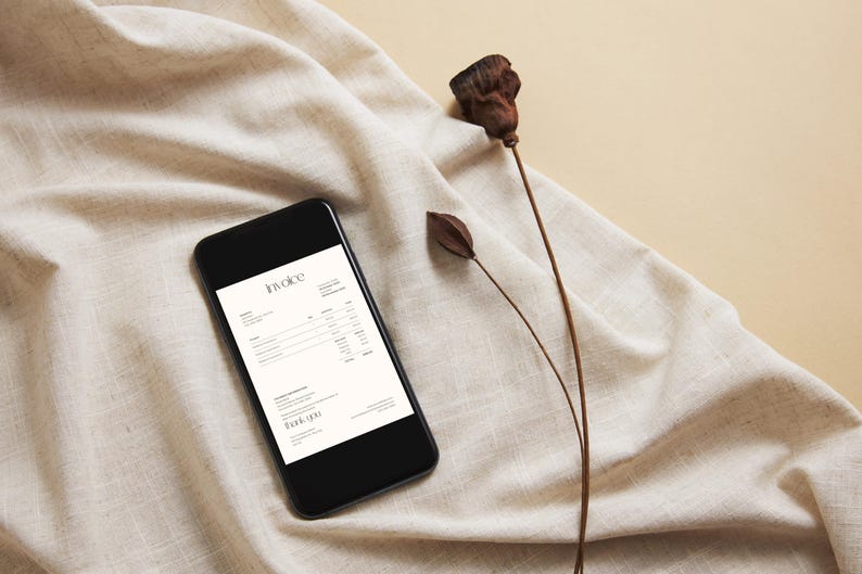 May include: A black smartphone displays an invoice on a cream-colored screen, resting on a beige linen cloth. Dried seed pods and stems add a natural element to the composition. The word "Invoice" is visible.