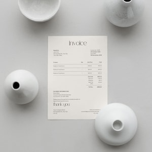 May include: An overhead shot features a cream-colored invoice with the word "invoice" at the top, surrounded by four white ceramic vases and a small bowl. The invoice includes details such as project names and amounts.