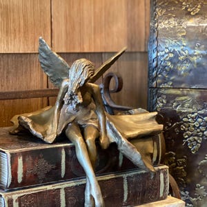 May include: A bronze-colored fairy sculpture with outstretched wings sits atop two stacked, antique-looking books. The fairy is draped in a flowing gown. A decorative chest with grape vine detailing is in the background.