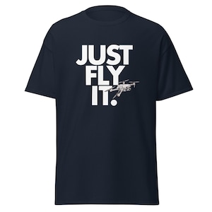 Könnte beinhalten: Marineblaues T-Shirt mit dem weißen Schriftzug "JUST FLY IT". Unter dem Text ist eine detaillierte Illustration einer Drohne abgebildet. Es ist ein Freizeitkleidungsstück.