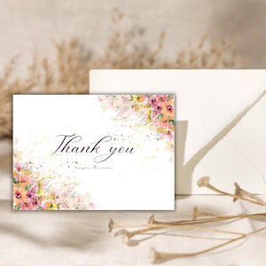 Peut inclure: Carte blanche avec l'inscription "Thank you" en écriture élégante, ornée de motifs floraux à l'aquarelle roses, jaunes et oranges. Une enveloppe est visible en arrière-plan, avec des éléments floraux séchés au premier plan.
