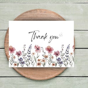 May include: A white card with the words "Thank you" in black script, adorned with watercolor flowers in shades of pink, purple, and yellow. The card is set on a wooden plate, creating a rustic aesthetic.