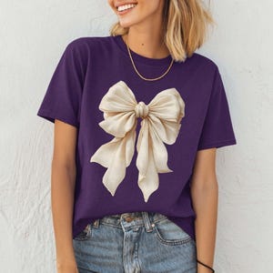 Peut inclure: Un t-shirt violet avec un grand n&oelig;ud de couleur cr&egrave;me. Le n&oelig;ud pr&eacute;sente des ombres d&eacute;taill&eacute;es, lui donnant un aspect r&eacute;aliste et en trois dimensions. Le t-shirt est de style col rond classique et est associ&eacute; &agrave; un jean bleu.