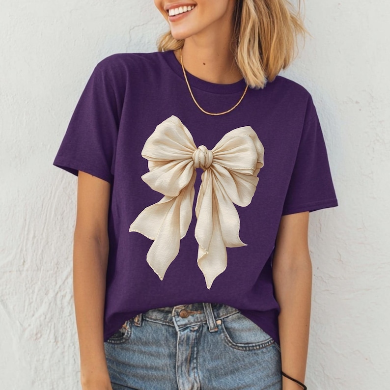 Peut inclure: Un t-shirt violet avec un grand n&oelig;ud de couleur cr&egrave;me. Le n&oelig;ud pr&eacute;sente des ombres d&eacute;taill&eacute;es, lui donnant un aspect r&eacute;aliste et en trois dimensions. Le t-shirt est de style col rond classique et est associ&eacute; &agrave; un jean bleu.