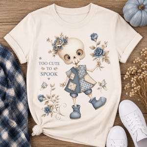 Peut inclure: T-shirt crème avec un personnage de squelette fantaisiste, des détails floraux bleus et des vêtements en patchwork. Le texte "TOO CUTE TO SPOOK" est imprimé. Des baskets blanches et une chemise à carreaux bleue sont visibles.