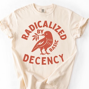 以下が含まれることがあります： クリーム色のTシャツに赤いグラフィック。グラフィックは小枝をくわえた鳥と「RADICALIZED BY BASIC DECENCY」という言葉を円形にデザインしています。Tシャツは半袖でクルーネックです。