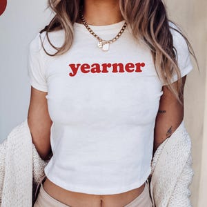 Puede incluir: Una camiseta blanca de manga corta con la palabra "yearner" impresa en letras minúsculas rojas en el pecho. La camiseta se lleva con un collar de cadena dorada y un cárdigan de punto color crema sobre los hombros.