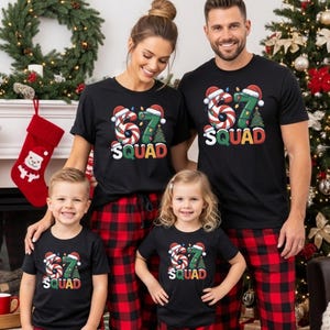 May include: A family of four wearing matching black t-shirts and red and black plaid pajama pants. The shirts have a colorful graphic with the text "67 SQUAD" and Christmas-themed elements. A Christmas tree and fireplace are in the background.