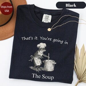 May include: Black t-shirt with the text "That's it. You're going in" and "The Soup". The graphic shows a frog wearing a chef's hat and apron, stirring a pot. A gold necklace is draped on the shirt.