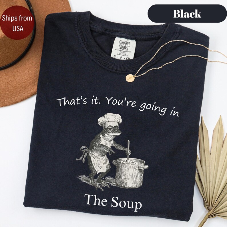 May include: Black t-shirt with the text "That's it. You're going in" and "The Soup". The graphic shows a frog wearing a chef's hat and apron, stirring a pot. A gold necklace is draped on the shirt.