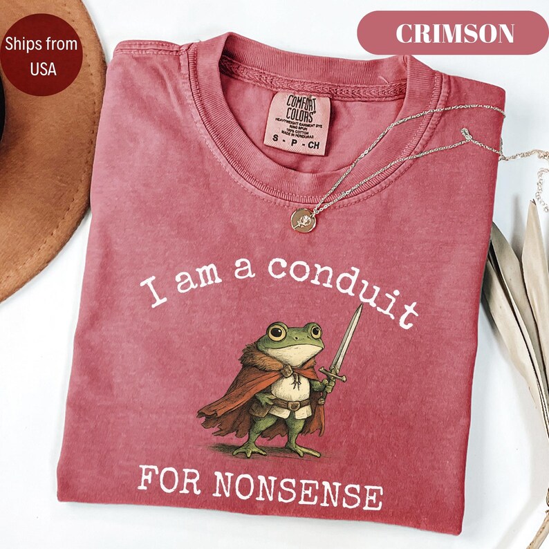 Peut inclure: T-shirt couleur cramoisi avec le texte "I am a conduit FOR NONSENSE" et un dessin de grenouille avec une &eacute;p&eacute;e. Le t-shirt a un col rond et des manches courtes. Un collier argent&eacute; avec un pendentif est pos&eacute; sur le t-shirt.