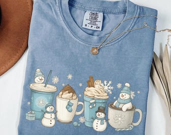 May include: Ice blue Comfort Colors t-shirt with a winter-themed graphic. The graphic features various mugs and cups of hot beverages, each with a snowman. A gold necklace is draped across the shirt.